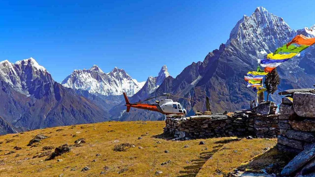 A red helicopter parked on a Himalayan ridge with prayer flags and snow-capped peaks.