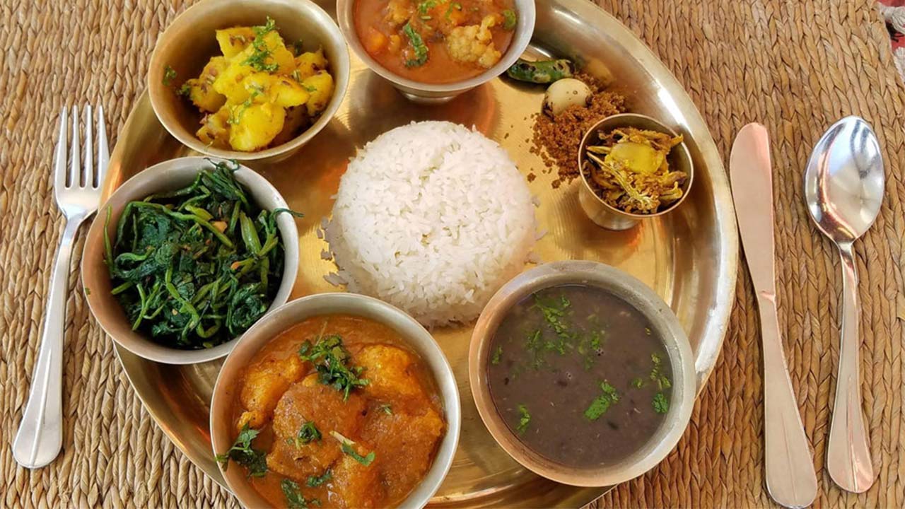 A top-down view of a traditional Nepali meal served on a metal platter, featuring a central mound of white rice surrounded by small bowls of dal, vegetable curry, saag (greens), and condiments with a spoon and fork on the side.
